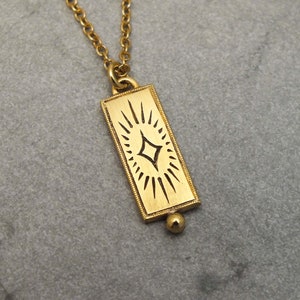 Może przedstawiać: Złoty naszyjnik z prostokątnym wisiorem w kształcie rozgwieżdżonego słońca. Wisiorek ma małą okrągłą perełkę na dole.