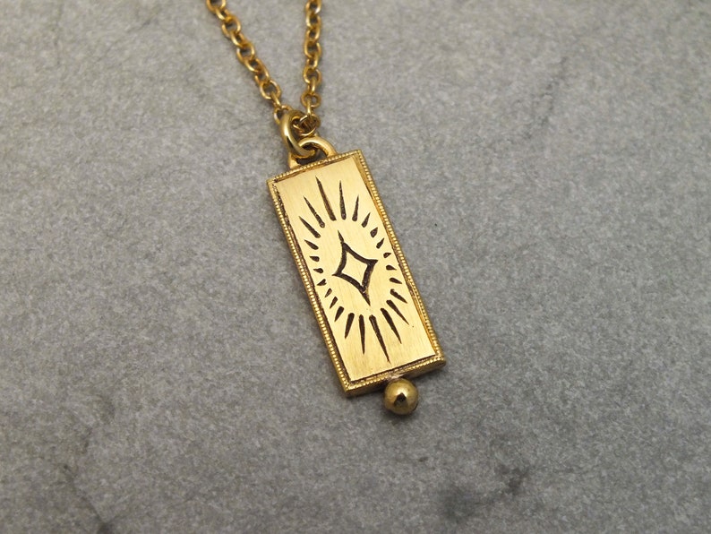 Może przedstawiać: Złoty naszyjnik z prostokątnym wisiorem w kształcie rozgwieżdżonego słońca. Wisiorek ma małą okrągłą perełkę na dole.