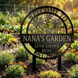 May include: Black metal garden sign with the words "WELCOME TO" in a circular design above a banner that reads "NANA'S GARDEN". The sign also includes the text "LOVE GROWS HERE" and "EST 2023". The sign features illustrations of vegetables.