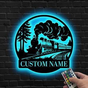 May include: Black metal train silhouette with a blue LED glow, set within a circular frame. The design features a steam engine, train cars, and the text "CUSTOM NAME" at the bottom. The image is mounted on a dark brick wall.