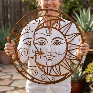 May include: A circular, rusty-brown metal wall art piece featuring a sun and moon design. The sun has a smiling face with rays, and the moon is depicted with a face and a star. The artwork is held up in front of a blurred background of plants.