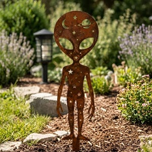 Op de afbeelding: Een roestkleurige metalen tuinsteker in de vorm van een alienfiguur. Het oppervlak is voorzien van uitgesneden ontwerpen van een halve maan, een planeet met ringen en diverse sterren verspreid over het hoofd en lichaam van het silhouet.