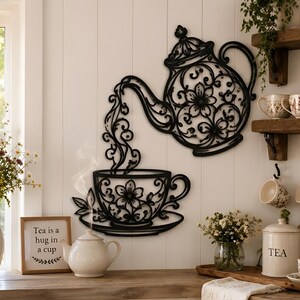 May include: A black metal wall art piece featuring a teapot pouring liquid into a teacup, both decorated with intricate floral cutouts. The design shows a teapot above a teacup with swirling patterns representing liquid flow, mounted on a white wall.