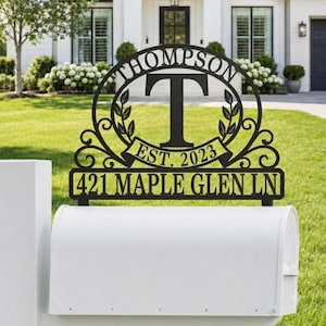 May include: A black metal mailbox topper featuring the name THOMPSON, a large letter T, the text EST. 2023, and the address 421 MAPLE GLEN LN. The design includes decorative scrollwork and leaf motifs, mounted securely atop a white mailbox.