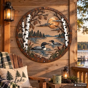 May include: A circular metal wall art piece featuring a lake scene with two loons swimming, birch trees, cattails, and a full moon. The design is framed in a rusted metal ring, depicting a serene nature landscape with layered silhouettes.