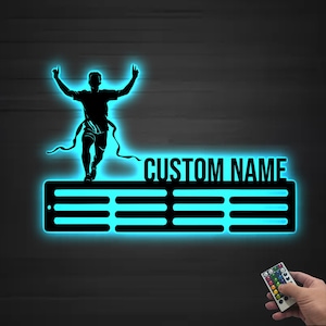Puede incluir: Un expositor de medallas de metal negro con una silueta de un corredor cruzando la línea de meta. La pantalla tiene un brillo azul y el texto "CUSTOM NAME" es visible. También se muestra un mando a distancia con varios botones de colores.