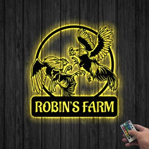 Puede incluir: Un letrero iluminado amarillo con dos gallos peleando dentro de un círculo. El letrero dice "ROBIN'S FARM" en un rectángulo negro debajo del círculo.