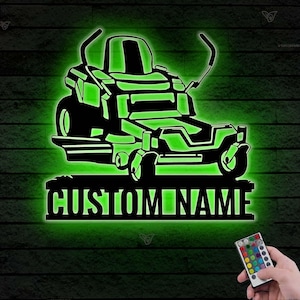 May include: A green illuminated silhouette of a ride-on lawnmower with the text "CUSTOM NAME" below. A remote control with multiple coloured buttons is visible in the bottom right corner.
