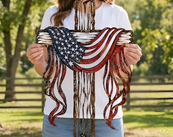 May include: A decorative metal cross wall hanging featuring a layered design with a distressed American flag pattern in red, white, and blue. The cross has a textured, weathered finish with flowing, ribbon-like metal strips draped across the center.