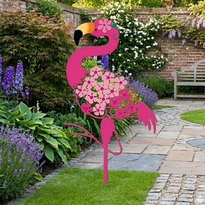 Può includere: Un fenicottero rosa in metallo per il giardino, con becco nero e dettagli gialli. Il fenicottero presenta un design floreale sul corpo e sulla testa, sullo sfondo di un muro di mattoni, piante e un sentiero di pietra.