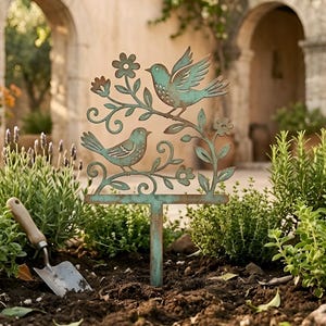 May include: A weathered metal garden stake featuring two birds perched on flowering branches. The stake has a T-shaped base for easy placement in soil. The birds and flowers have a verdigris finish, adding a touch of vintage charm to any garden.