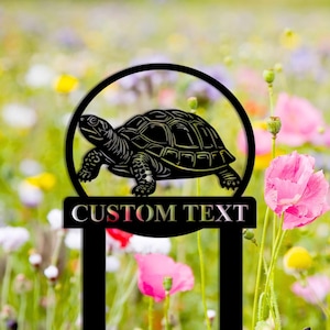 Op de afbeelding: Zwarte metalen tuinstek met een silhouet van een schildpad en de tekst "CUSTOM TEXT". De stek staat in een veld met wilde bloemen.