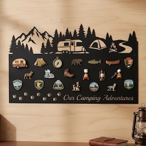Könnte beinhalten: Eine schwarze Metall-Anzeigetafel mit einer Berg- und Waldsilhouette oben und dem Text Our Camping Adventures unten. Die Tafel ist mit verschiedenen Magneten zum Thema Camping verziert, darunter ein Wohnwagen, ein Zelt, ein Kompass und Aufnäher.