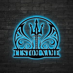 May include: Black metal wall art with a blue neon glow. The design features a trident at the center, surrounded by decorative swirls and a circular frame. The text "CUSTOM NAME" is displayed at the bottom, perfect for personalized decor.