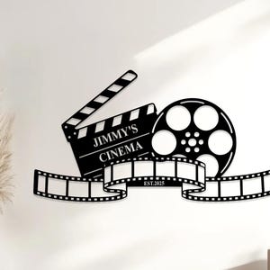 Könnte beinhalten: Schwarze Metall-Wandkunst mit einem Filmklappbrett, einer Filmrolle und einem Filmstreifen-Design. Auf dem Klappbrett steht "JIMMY'S CINEMA" und "EST.2025". Ein dekoratives Stück für ein Heimkino oder einen Kinoraum.