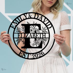 May include: Black metal circular wall art with the names "Emily & David" and "Elizabeth" and the year "EST 2026". The design features floral and leaf accents around the letter "E".