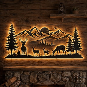 Puede incluir: Una pieza de arte mural de metal negro que muestra un paisaje montañoso con un sol grande, dos pinos altos y una familia de cuatro ciervos. El diseño de silueta está retroiluminado con luz cálida, creando un efecto brillante en la pared.