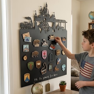 Puede incluir: Un tablero de pared negro con una silueta cortada a láser de un castillo, un tren y una brújula en la parte superior. El tablero incluye el texto "Oh, the places I've been..." y está decorado con varios imanes pequeños y coloridos de temática viajera.