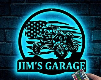 May include: A black metal sign with a blue LED light, featuring an off-road vehicle silhouette and an American flag design. The sign reads "JIM'S GARAGE" in white letters. A hand holds a remote control.