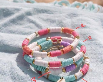 May include: Four colorful beaded bracelets with gold accents and a small seashell charm. The bracelets are stacked on top of each other and are made of light blue, white, pink, and purple beads.