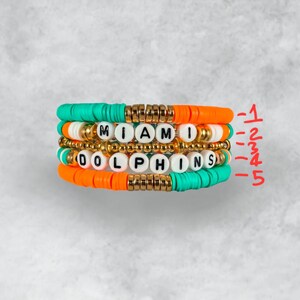 May include: A set of four beaded bracelets with the words "Miami Dolphins" spelled out in white letters. The bracelets are made with orange, turquoise, and gold beads.