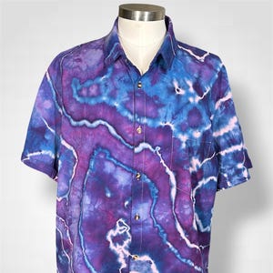 May include: A short-sleeve button-down shirt featuring a vibrant tie-dye pattern in shades of deep purple, royal blue, and bright pink. The shirt has a classic collar, a front button placket with visible buttons, and a single chest pocket on the left side.
