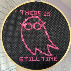 May include: A cross-stitch hoop featuring a black background with pink text and a ghost-like figure wearing glasses. The text reads "THERE IS STILL TIME". The hoop is held in a wooden frame.