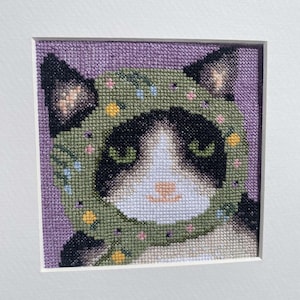 May include: A black and white cat wearing a green and floral hooded hat. The cat's eyes are green and the hat has a white trim.