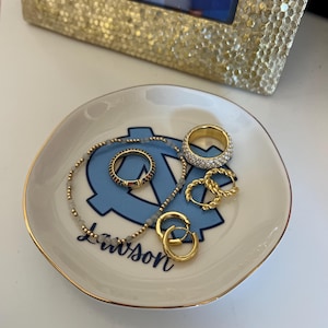 Puede incluir: Un peque&ntilde;o plato de baratijas blanquecino con un borde dorado y un logotipo azul claro. El plato contiene varios anillos dorados, un collar de cuentas y pendientes de aro dorados. Una foto con marco dorado est&aacute; en el fondo.