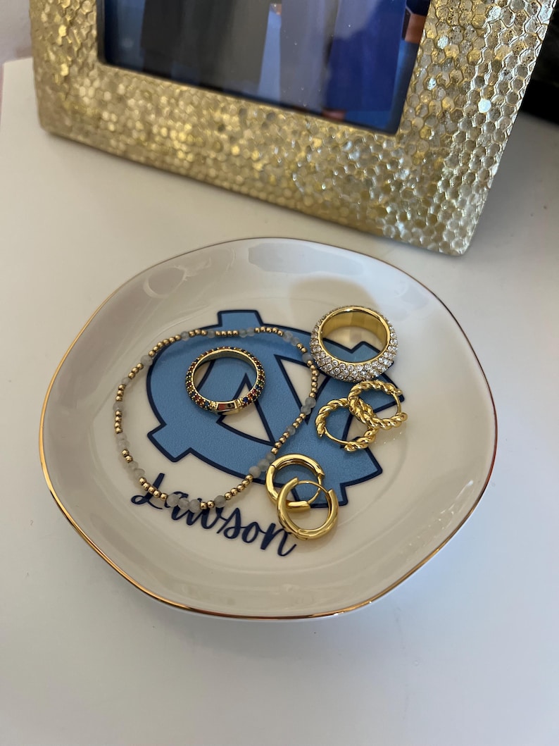 Puede incluir: Un peque&ntilde;o plato de baratijas blanquecino con un borde dorado y un logotipo azul claro. El plato contiene varios anillos dorados, un collar de cuentas y pendientes de aro dorados. Una foto con marco dorado est&aacute; en el fondo.