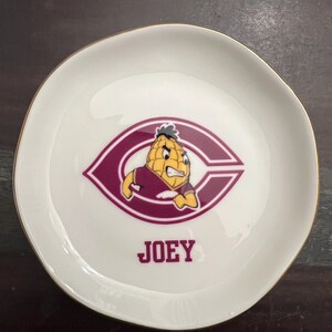 Puede incluir: Un plato de cer&aacute;mica blanco con borde dorado. El plato presenta un logotipo granate y blanco con un personaje de dibujos animados de ma&iacute;z y el nombre "JOEY" en texto granate.