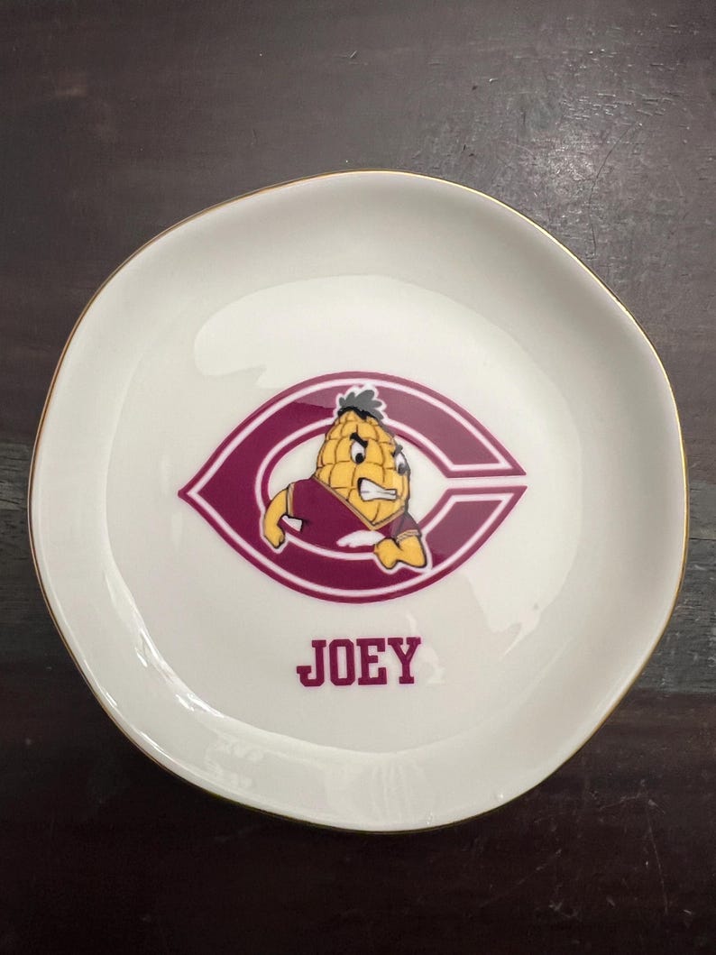 Puede incluir: Un plato de cer&aacute;mica blanco con borde dorado. El plato presenta un logotipo granate y blanco con un personaje de dibujos animados de ma&iacute;z y el nombre "JOEY" en texto granate.