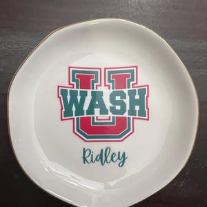 Puede incluir: Plato de cer&aacute;mica blanco con borde dorado. El plato presenta un dise&ntilde;o verde y rojo con la palabra "WASH" y la letra "U". El nombre "Ridley" est&aacute; escrito debajo en letra cursiva.