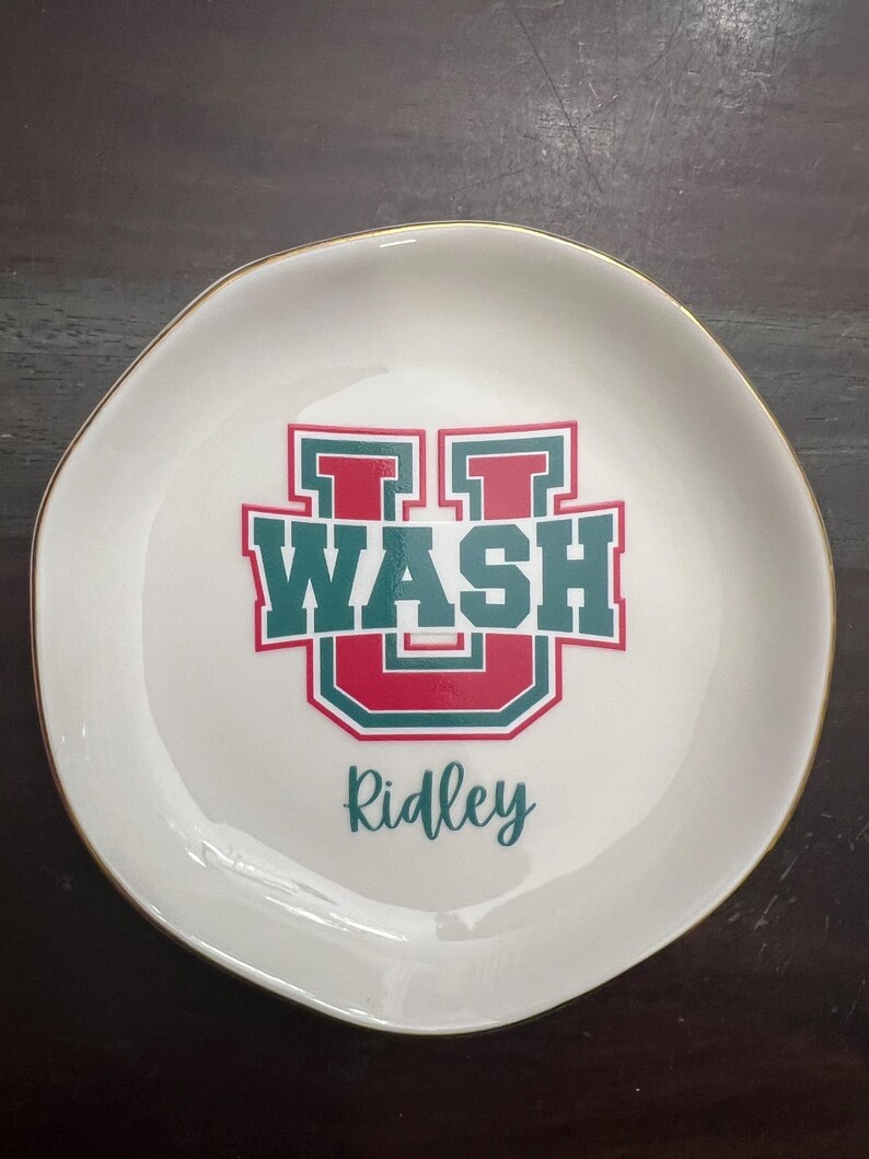 Puede incluir: Plato de cer&aacute;mica blanco con borde dorado. El plato presenta un dise&ntilde;o verde y rojo con la palabra "WASH" y la letra "U". El nombre "Ridley" est&aacute; escrito debajo en letra cursiva.
