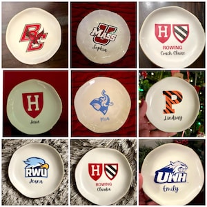 Puede incluir: Nueve platos de cer&aacute;mica blanquecina con bordes dorados, cada uno con un logotipo o inicial universitario diferente. Algunos platos tienen nombres debajo de los logotipos. Los logotipos incluyen un &aacute;guila roja, un elefante azul y una P con rayas de tigre.