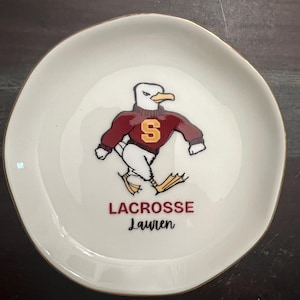 Puede incluir: Un plato de cer&aacute;mica blanco con borde dorado. El plato presenta una gaviota de dibujos animados con un su&eacute;ter granate con una "S" dorada. Debajo, las palabras "LACROSSE Lauren" est&aacute;n impresas en granate.