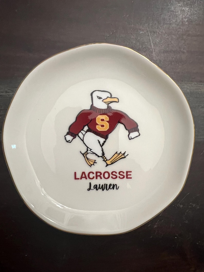 Puede incluir: Un plato de cer&aacute;mica blanco con borde dorado. El plato presenta una gaviota de dibujos animados con un su&eacute;ter granate con una "S" dorada. Debajo, las palabras "LACROSSE Lauren" est&aacute;n impresas en granate.