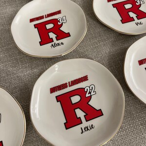 Puede incluir: Platos de cer&aacute;mica blanca para baratijas con bordes dorados. Cada plato presenta el texto "Rutgers Lacrosse" sobre una gran "R" roja con un contorno negro, un n&uacute;mero y un nombre. Los platos est&aacute;n personalizados.