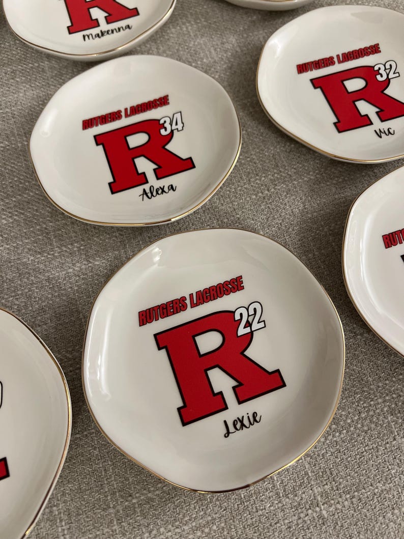 Puede incluir: Platos de cer&aacute;mica blanca para baratijas con bordes dorados. Cada plato presenta el texto "Rutgers Lacrosse" sobre una gran "R" roja con un contorno negro, un n&uacute;mero y un nombre. Los platos est&aacute;n personalizados.