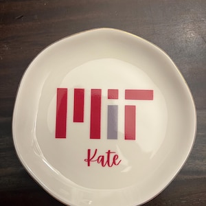 Puede incluir: Un plato de cer&aacute;mica blanco con borde dorado. El plato presenta las letras "MIT" en rojo y gris, con el nombre "Kate" en cursiva debajo. El plato es redondo con un borde ligeramente irregular.
