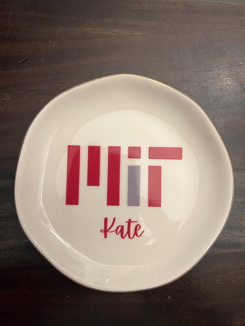 Puede incluir: Un plato de cer&aacute;mica blanco con borde dorado. El plato presenta las letras "MIT" en rojo y gris, con el nombre "Kate" en cursiva debajo. El plato es redondo con un borde ligeramente irregular.
