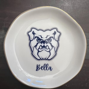 Puede incluir: Plato de cer&aacute;mica blanco para baratijas con borde dorado. El plato presenta un gr&aacute;fico de bulldog azul marino y el nombre "Bella" en escritura cursiva. El plato tiene una forma ligeramente irregular.
