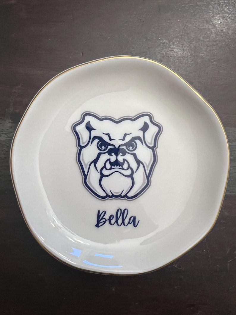 Puede incluir: Plato de cer&aacute;mica blanco para baratijas con borde dorado. El plato presenta un gr&aacute;fico de bulldog azul marino y el nombre "Bella" en escritura cursiva. El plato tiene una forma ligeramente irregular.