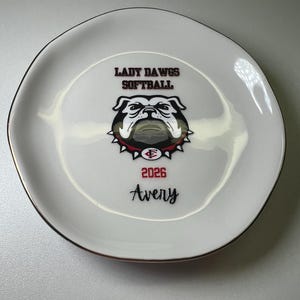 Puede incluir: Reposa cucharas de cer&aacute;mica blanca con un dise&ntilde;o negro y rojo. El dise&ntilde;o incluye el texto "LADY DAWGS SOFTBALL", un gr&aacute;fico de bulldog, el a&ntilde;o "2026" y el nombre "Avery". El reposa cucharas tiene un borde dorado.