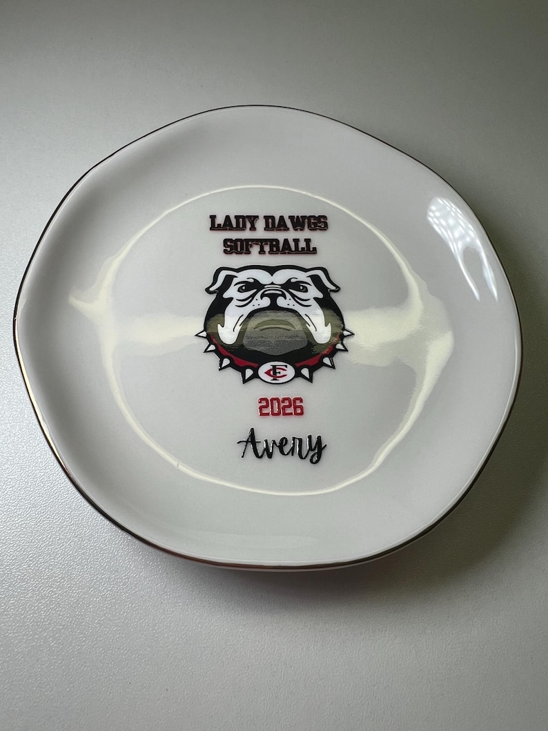 Puede incluir: Reposa cucharas de cer&aacute;mica blanca con un dise&ntilde;o negro y rojo. El dise&ntilde;o incluye el texto "LADY DAWGS SOFTBALL", un gr&aacute;fico de bulldog, el a&ntilde;o "2026" y el nombre "Avery". El reposa cucharas tiene un borde dorado.