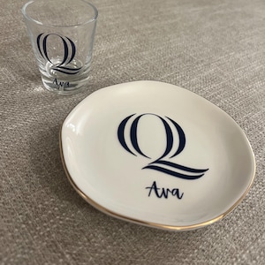 Puede incluir: Un vaso de chupito transparente y un plato peque&ntilde;o blanco con borde dorado. Ambos art&iacute;culos presentan la letra "Q" estilizada en azul marino y el nombre "Ava". El plato es de forma ovalada.