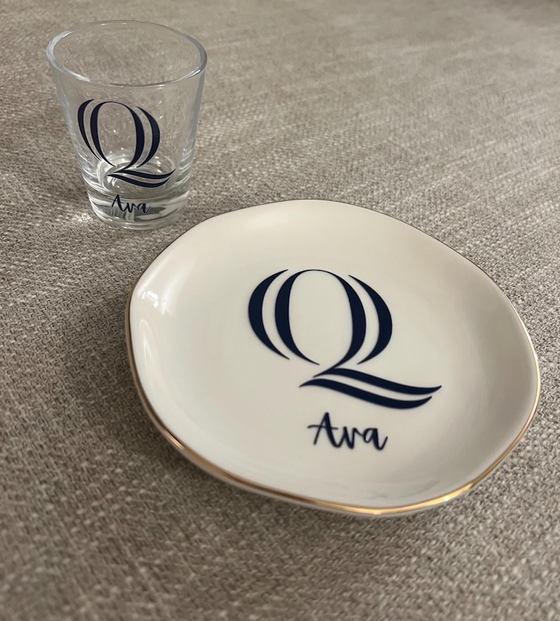 Puede incluir: Un vaso de chupito transparente y un plato peque&ntilde;o blanco con borde dorado. Ambos art&iacute;culos presentan la letra "Q" estilizada en azul marino y el nombre "Ava". El plato es de forma ovalada.