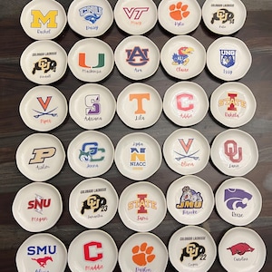 Puede incluir: Una colecci&oacute;n de peque&ntilde;os platos redondos de cer&aacute;mica blanca. Cada plato presenta un logotipo o mascota universitaria diferente, junto con un nombre. Los logotipos son de varios colores, incluyendo naranja, morado y azul. Los nombres est&aacute;n escritos en una fuente cursiva.