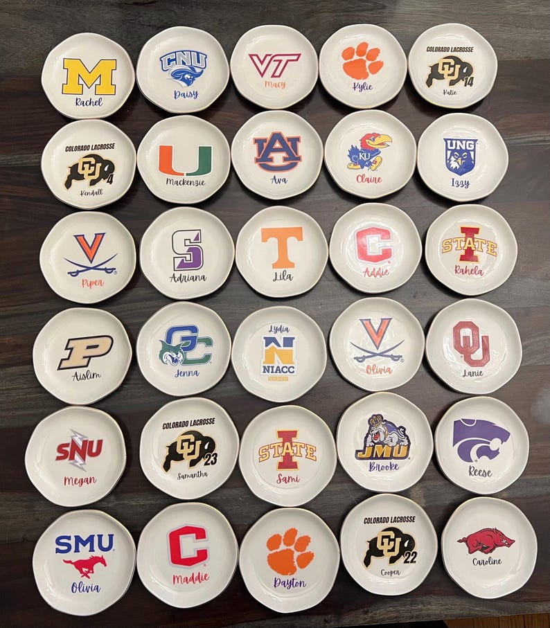 Puede incluir: Una colecci&oacute;n de peque&ntilde;os platos redondos de cer&aacute;mica blanca. Cada plato presenta un logotipo o mascota universitaria diferente, junto con un nombre. Los logotipos son de varios colores, incluyendo naranja, morado y azul. Los nombres est&aacute;n escritos en una fuente cursiva.