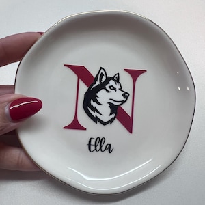 Puede incluir: Platito de cer&aacute;mica blanca con borde dorado. Presenta la letra "N" en granate con un gr&aacute;fico de perro husky en blanco y negro. El nombre "Ella" est&aacute; escrito en cursiva negra debajo.